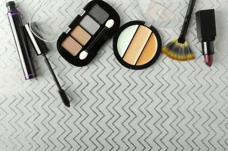 Cosmetics products including mascara, eyeshadow palette, concealer, fan brush, and lipstick on a patterned background, relevant to the cosmetics and chemicals sector in business law.